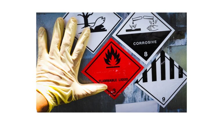 A gloved hand near hazardous waste labels, including signs for flammable liquid, corrosive material, and other dangerous substances.