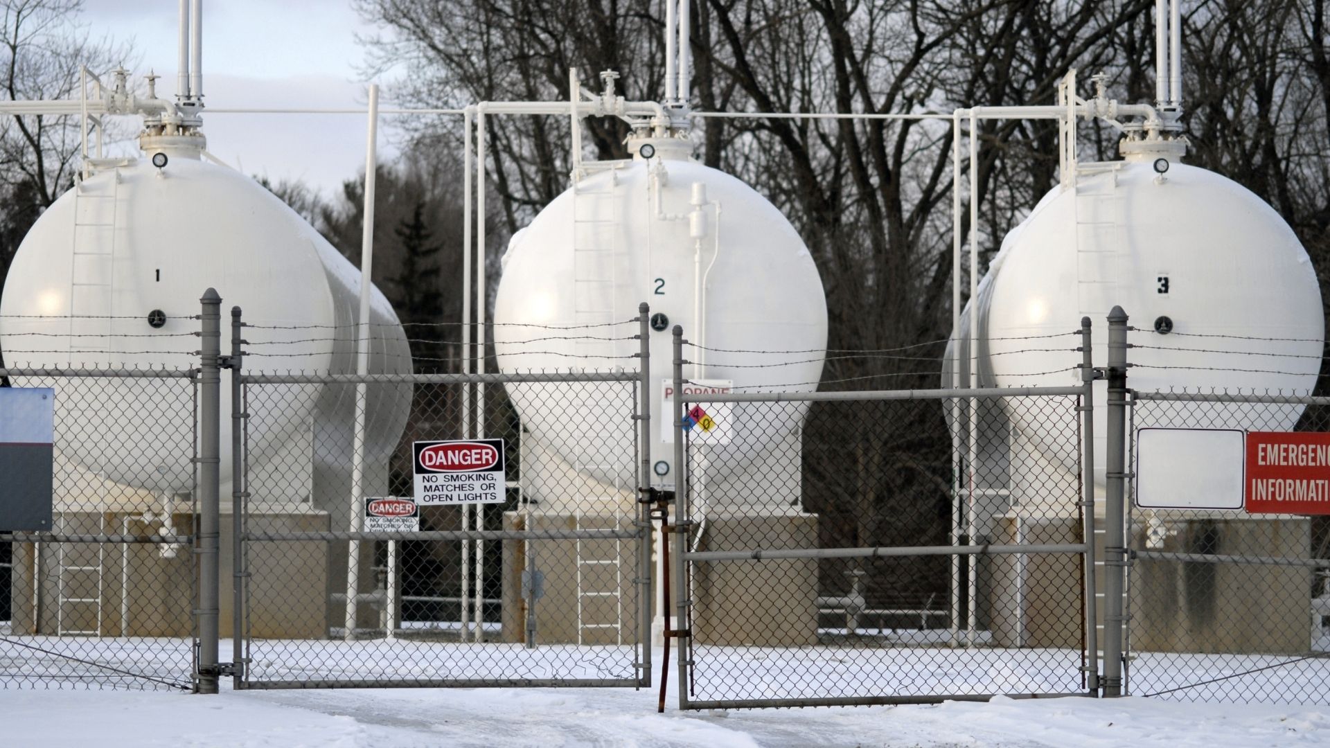 Large white propane storage tanks behind a fenced area with "DANGER" and "EMERGENCY INFORMATION" signs, surrounded by snow and trees.