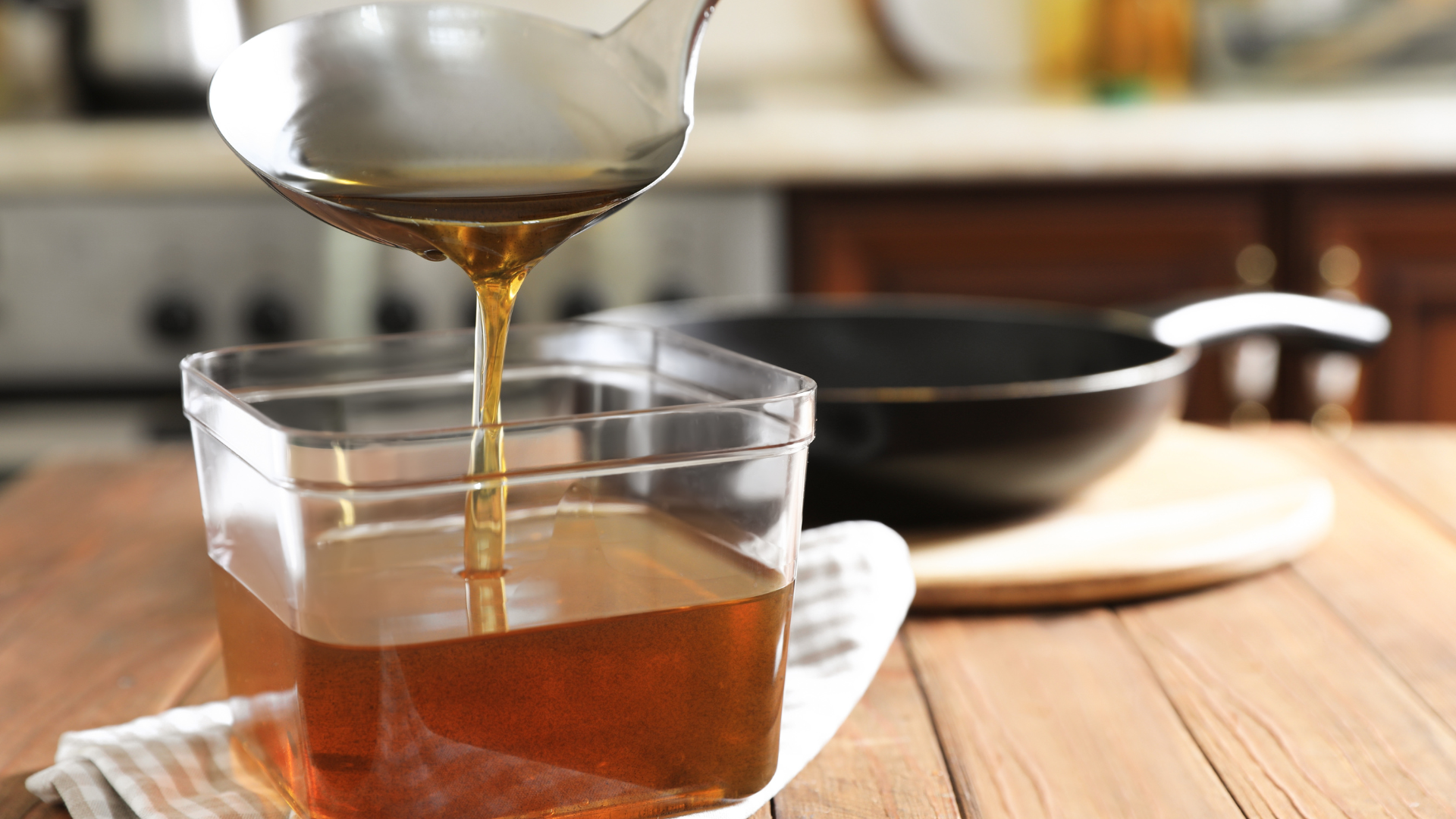 A ladle pouring golden oil into a clear container on a wooden table, with a black pan in the background, in a kitchen setting.