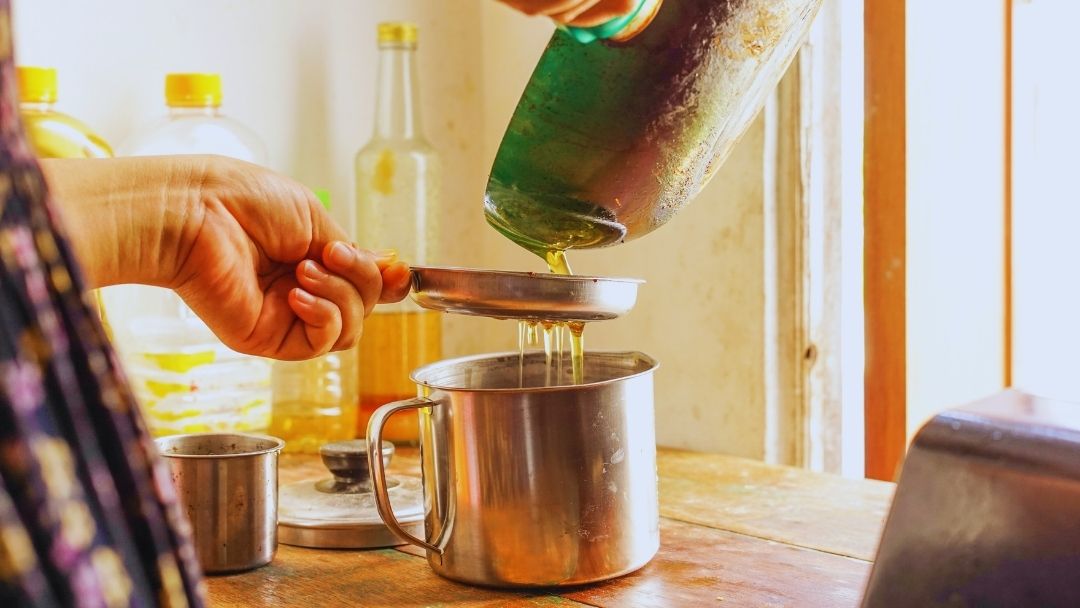 Person filtering used cooking oil through a strainer into a metal container, with oil storage bottles in the background for recycling.