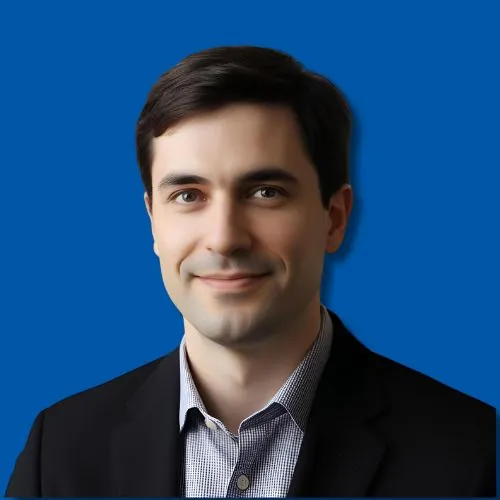 Headshot of smiling man with dark hair in dark blazer and checked shirt against blue background